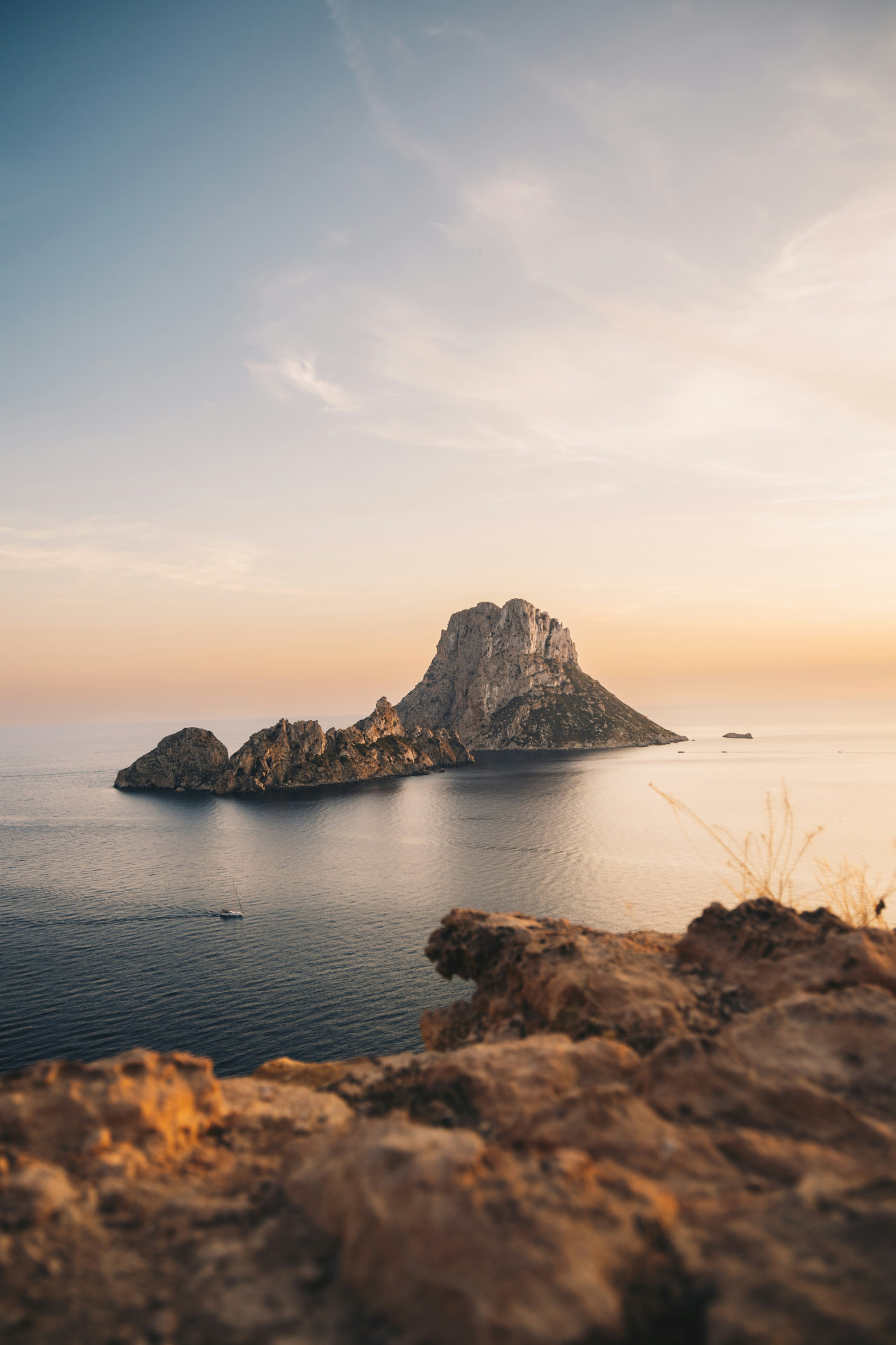 Es Vedrà rising from the sea at golden hour, seen from a rocky cliff viewpoint in Ibiza with a small boat on the water